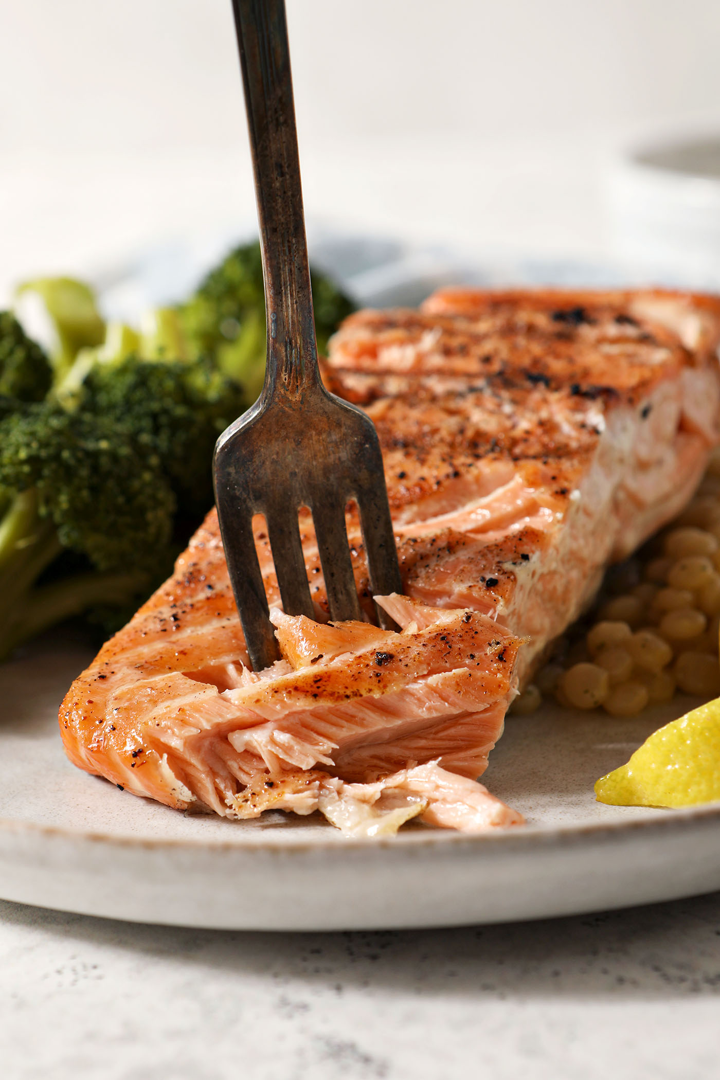 A fork slides into a filet of grilled salmon on a plate with broccoli and cous cous