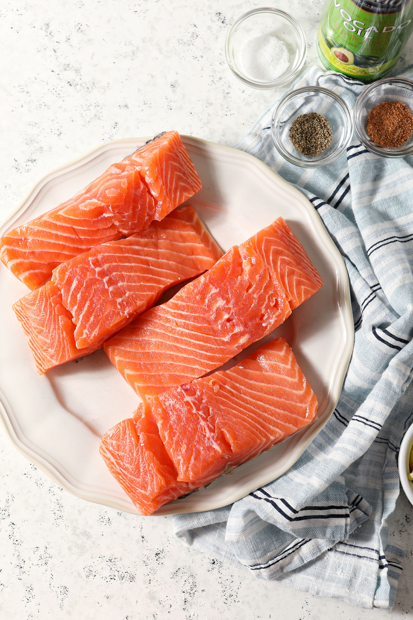 Overhead of salmon filets on a white plate on a stone surface surrounded with bowls of spices and a can of avocado oil spray