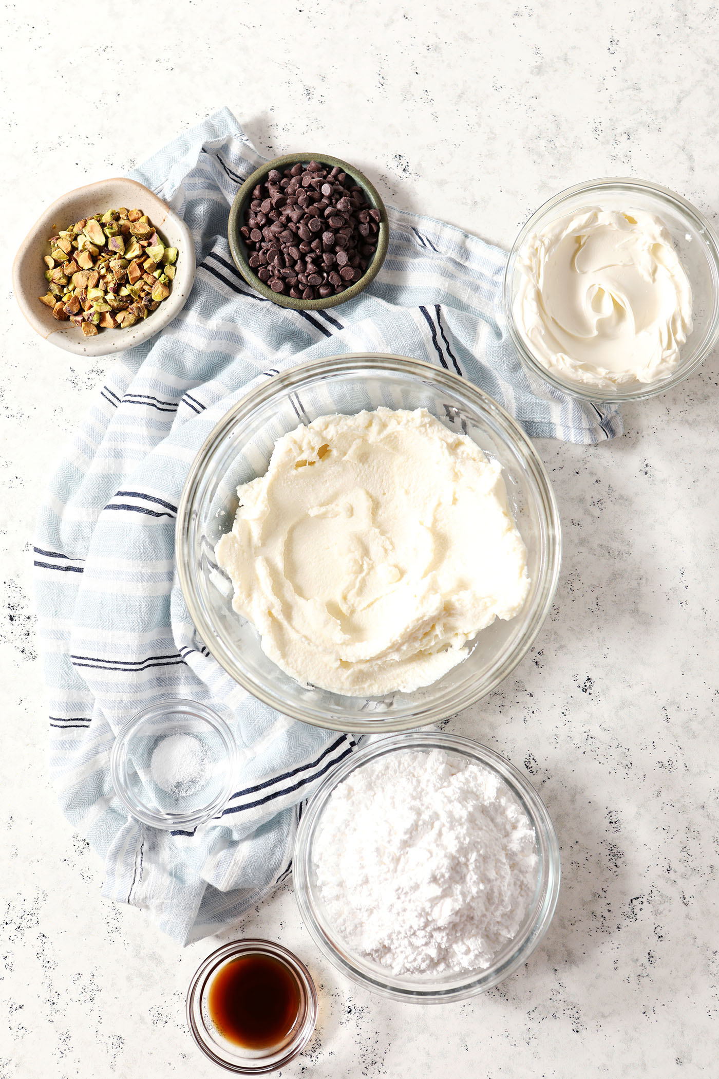 Ingredients to make a sweet Cannoli Dip in bowls on a light-colored stone surface with a blue striped linen