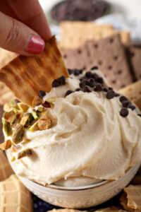 A piece of waffle cone dips into a bowl of Sweet Cannoli Dip garnish with chopped pistachios and chocolate chips.