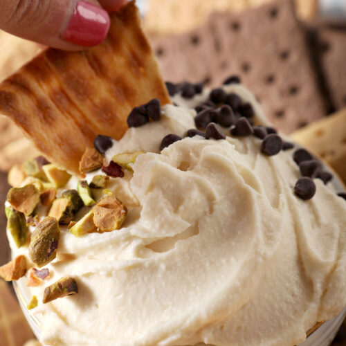 A piece of waffle cone dips into a bowl of Sweet Cannoli Dip garnish with chopped pistachios and chocolate chips.