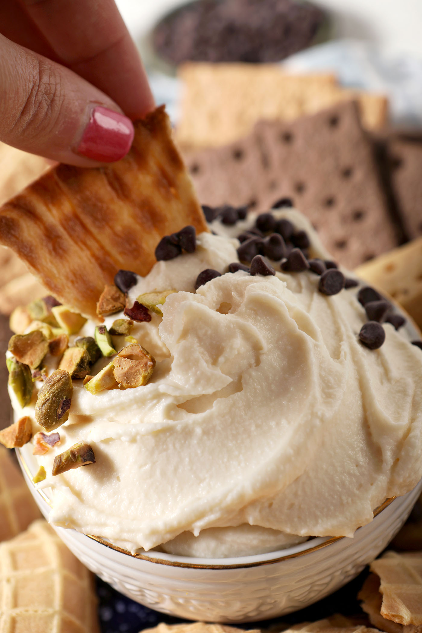 A piece of waffle cone dips into a bowl of Sweet Cannoli Dip garnish with chopped pistachios and chocolate chips.