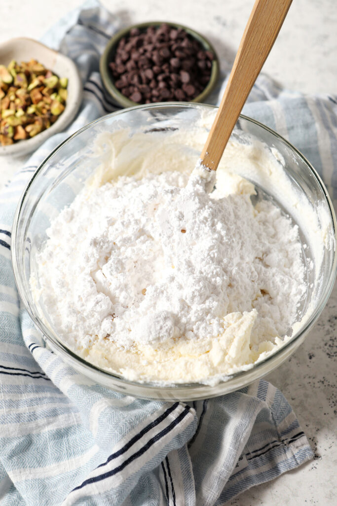 Powdered sugar sits on top of a ricotta mixture in a bowl with a rubber spatula on a stone surface with a blue striped linen