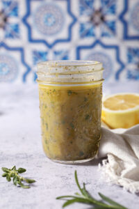 A mason jar of lemon vinaigrette on a stone surface in front of a blue and white tiled backsplash that reads "Lemon Vinaigrette"
