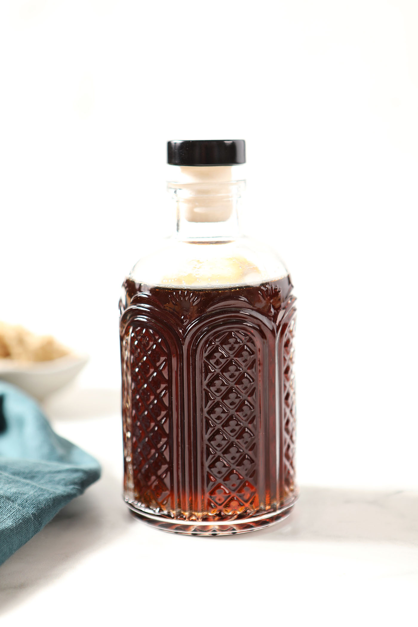 A bottle of brown sugar simple syrup on a white stone surface next to a turquoise linen and a bowl of brown sugar