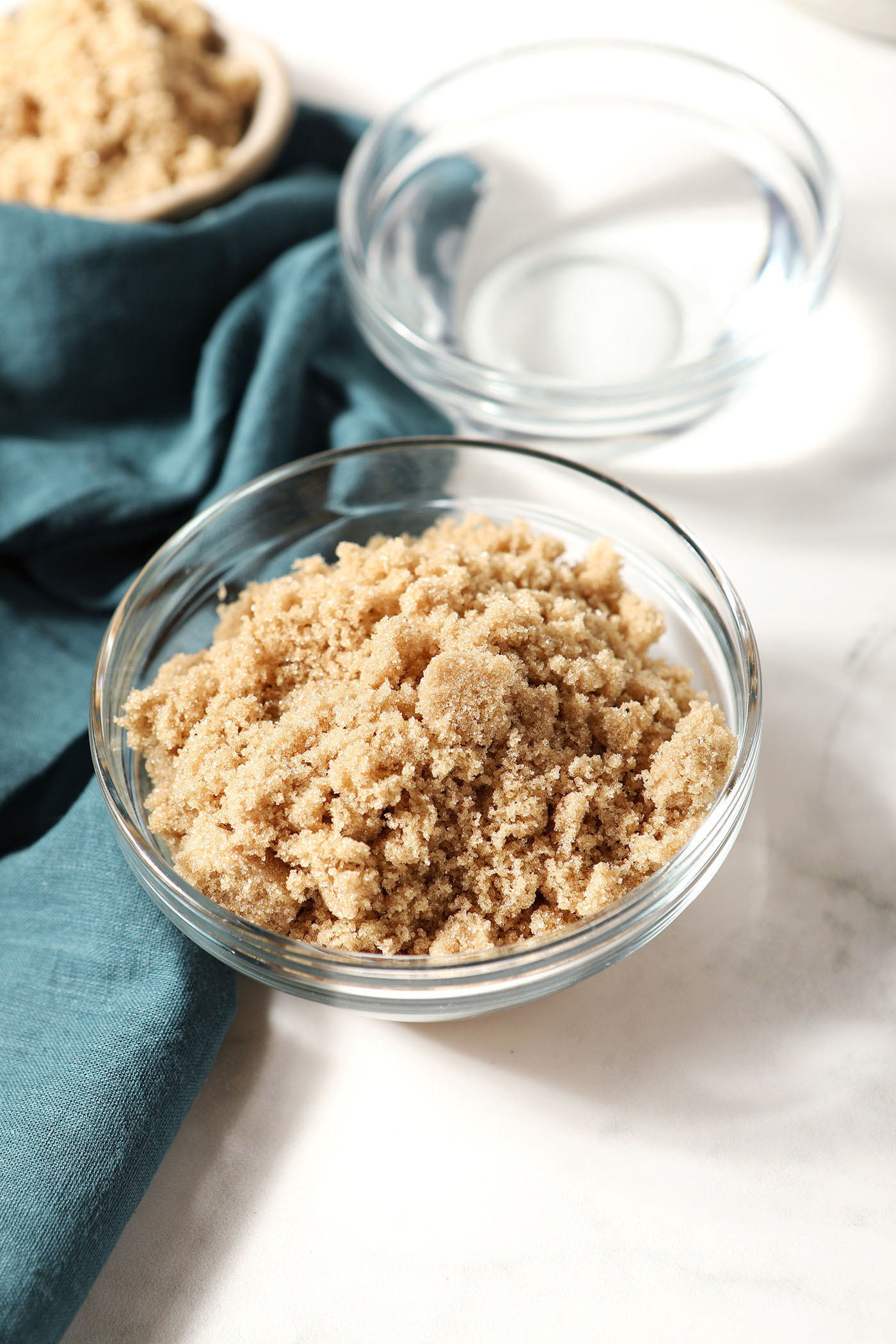 A bowl of brown sugar next to a bowl of water on a white stone surface with a turquoise linen