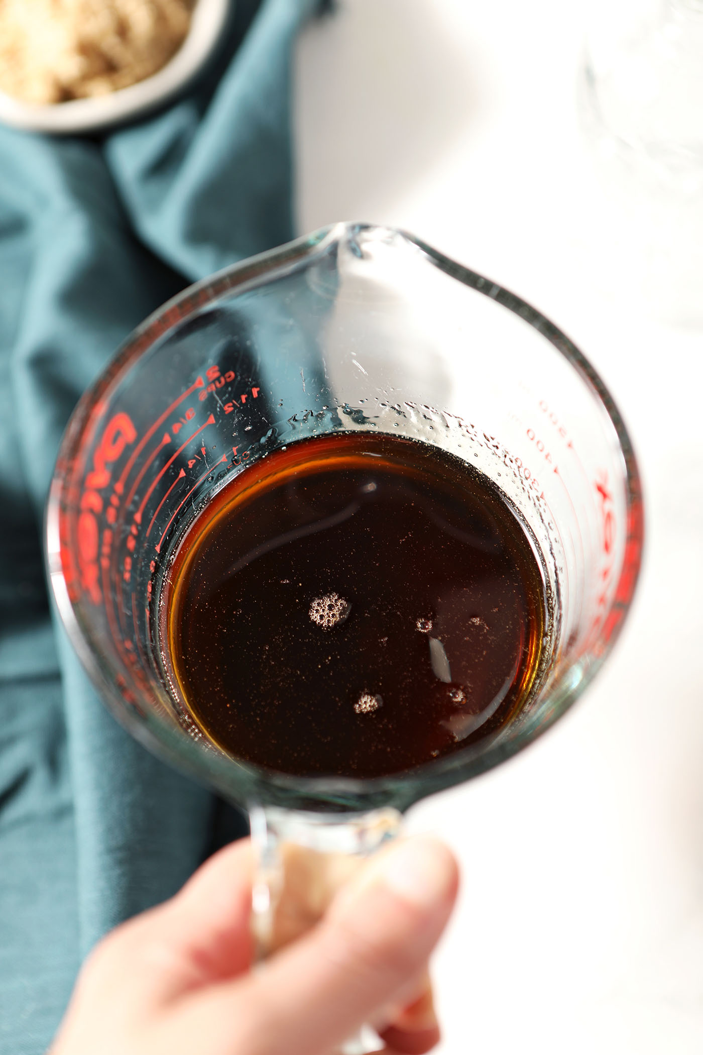 A hand holds a glass measuring cup of brown sugar syrup above a white stone surface and dark turquoise linen