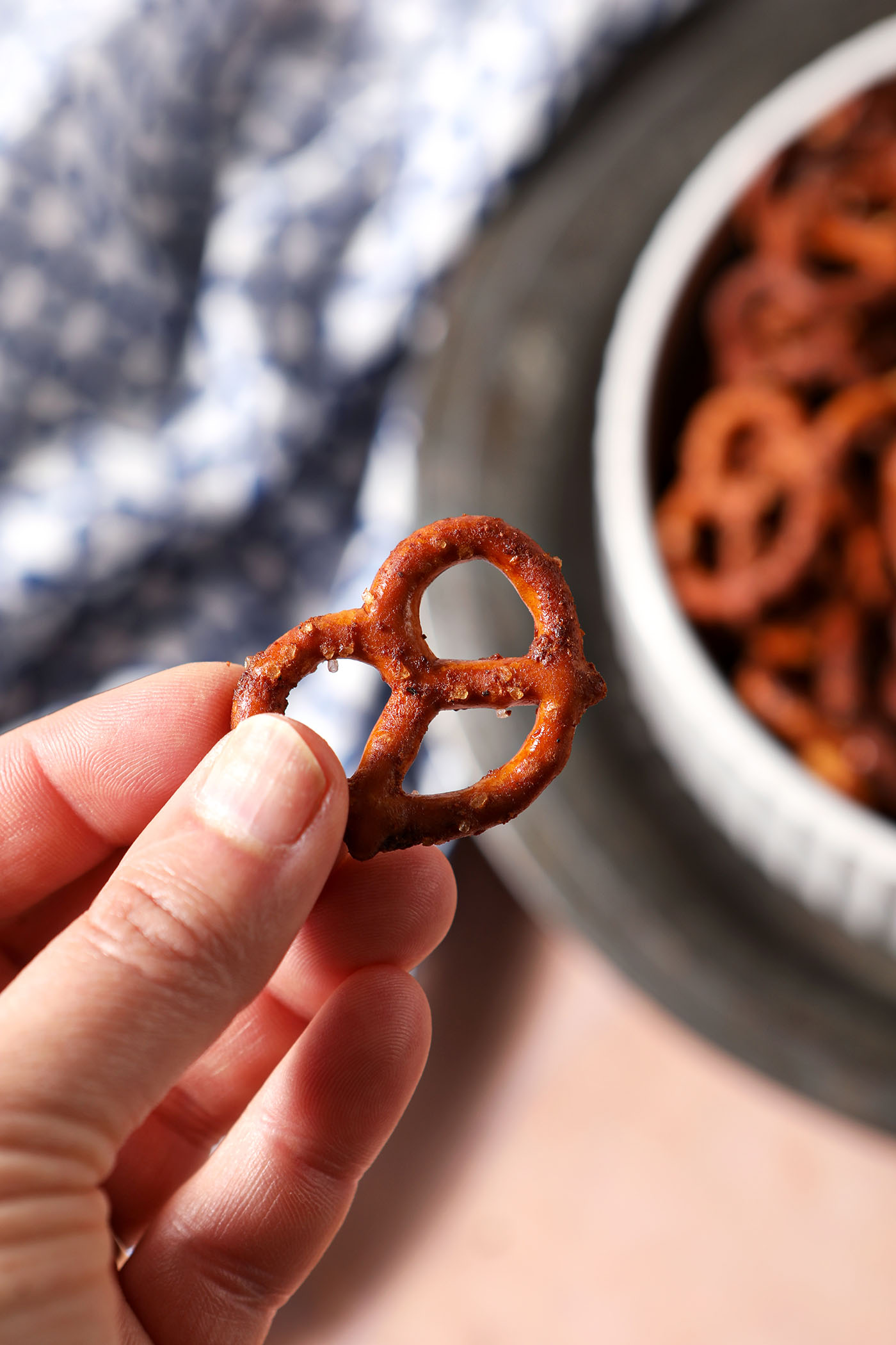 A hand holds a Cajun spice pretzel above a tan surface with a patterned blue and white linen and a bowl of pretzels