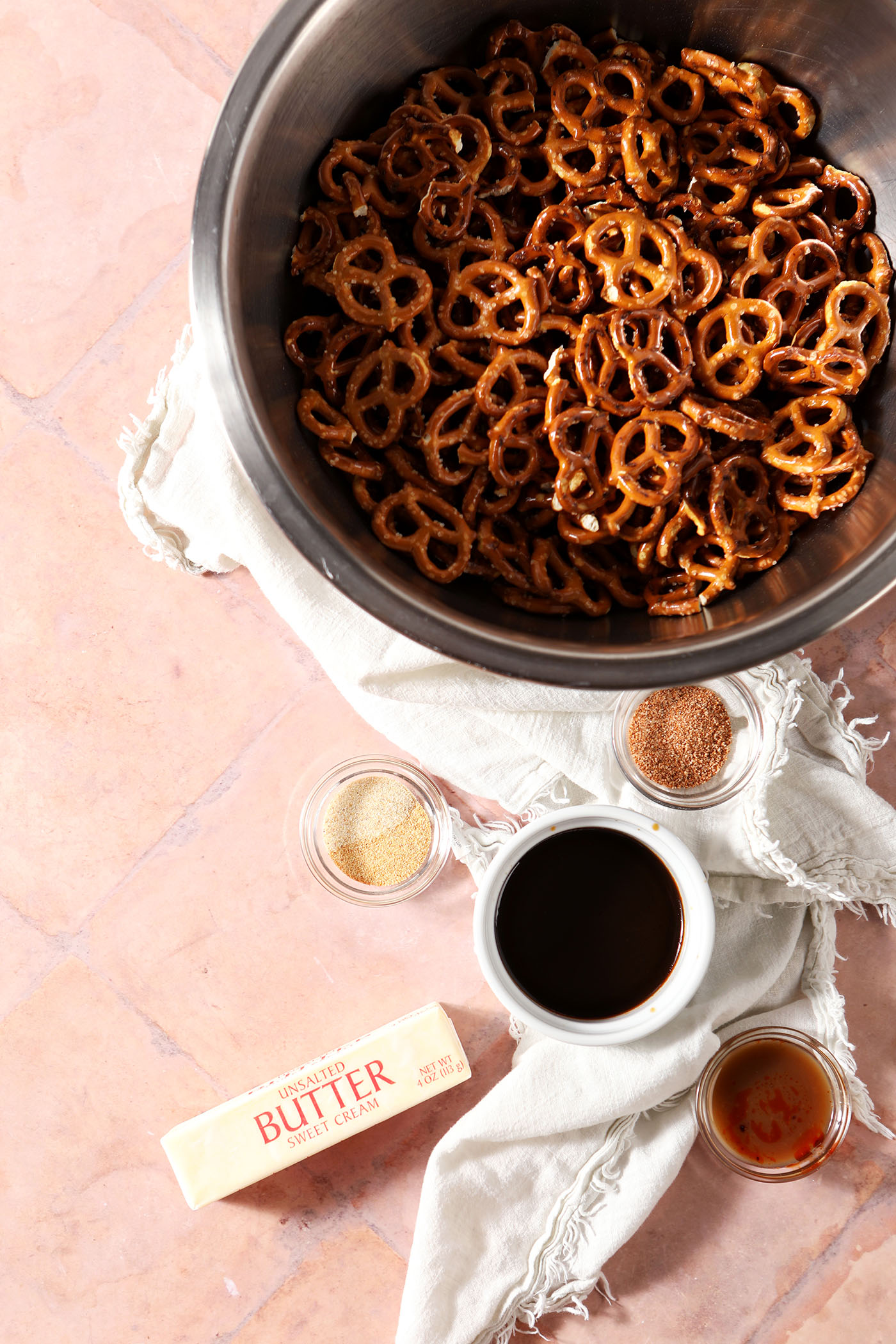 A bowl of crunchy pretzels sits next to small bowls of other ingredients and a stick of butter on a brick surface
