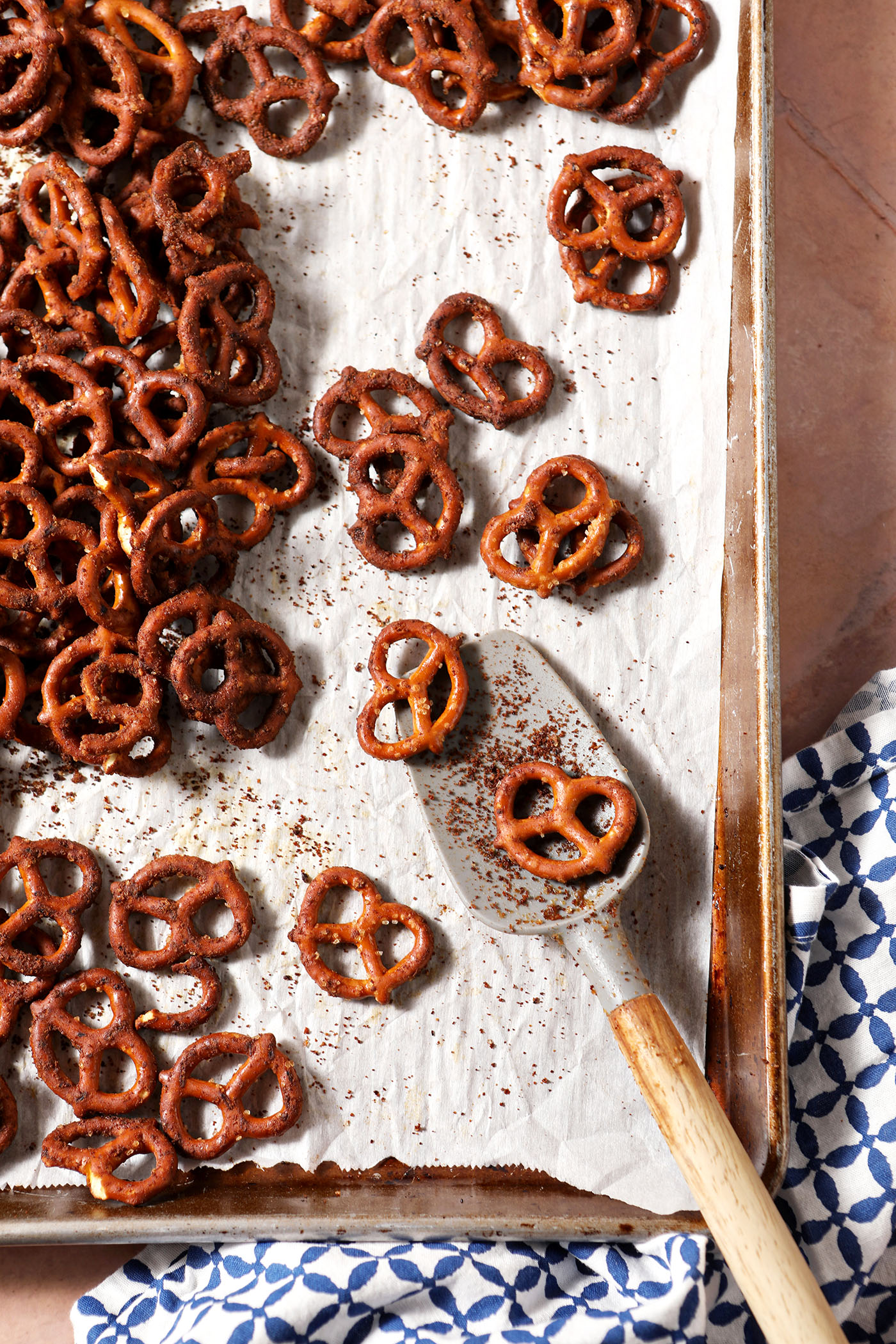 A spatula sits on a sheet pan of Cajun spice pretzels on a brick surface with a blue and white linen