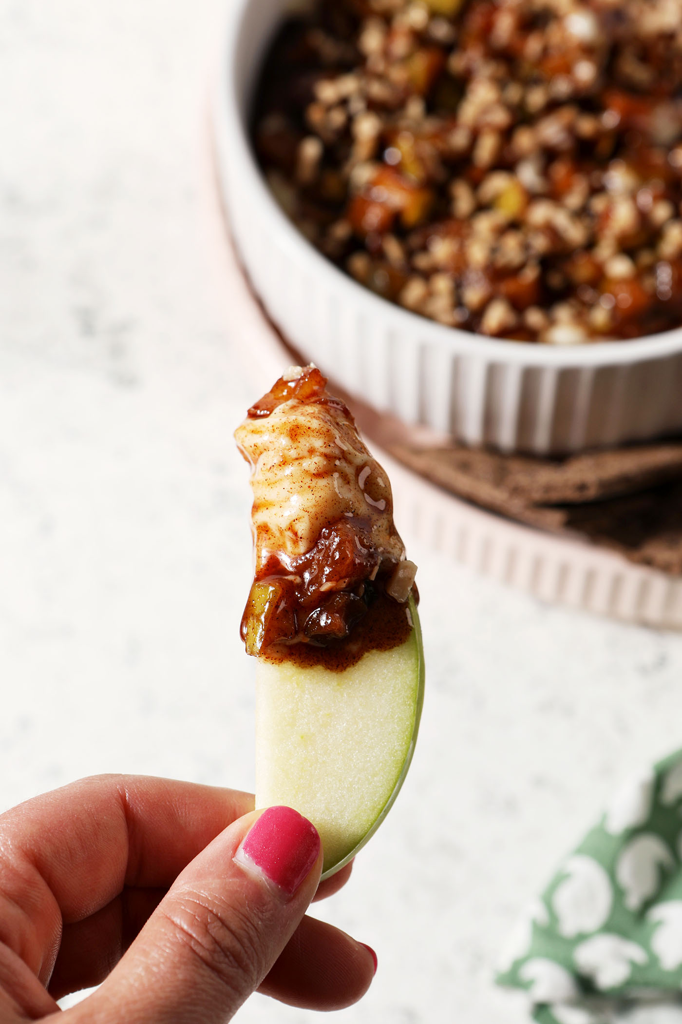 A hand holds an apple slice, holding Caramel Apple Cheesecake Dip above a platter with more dip