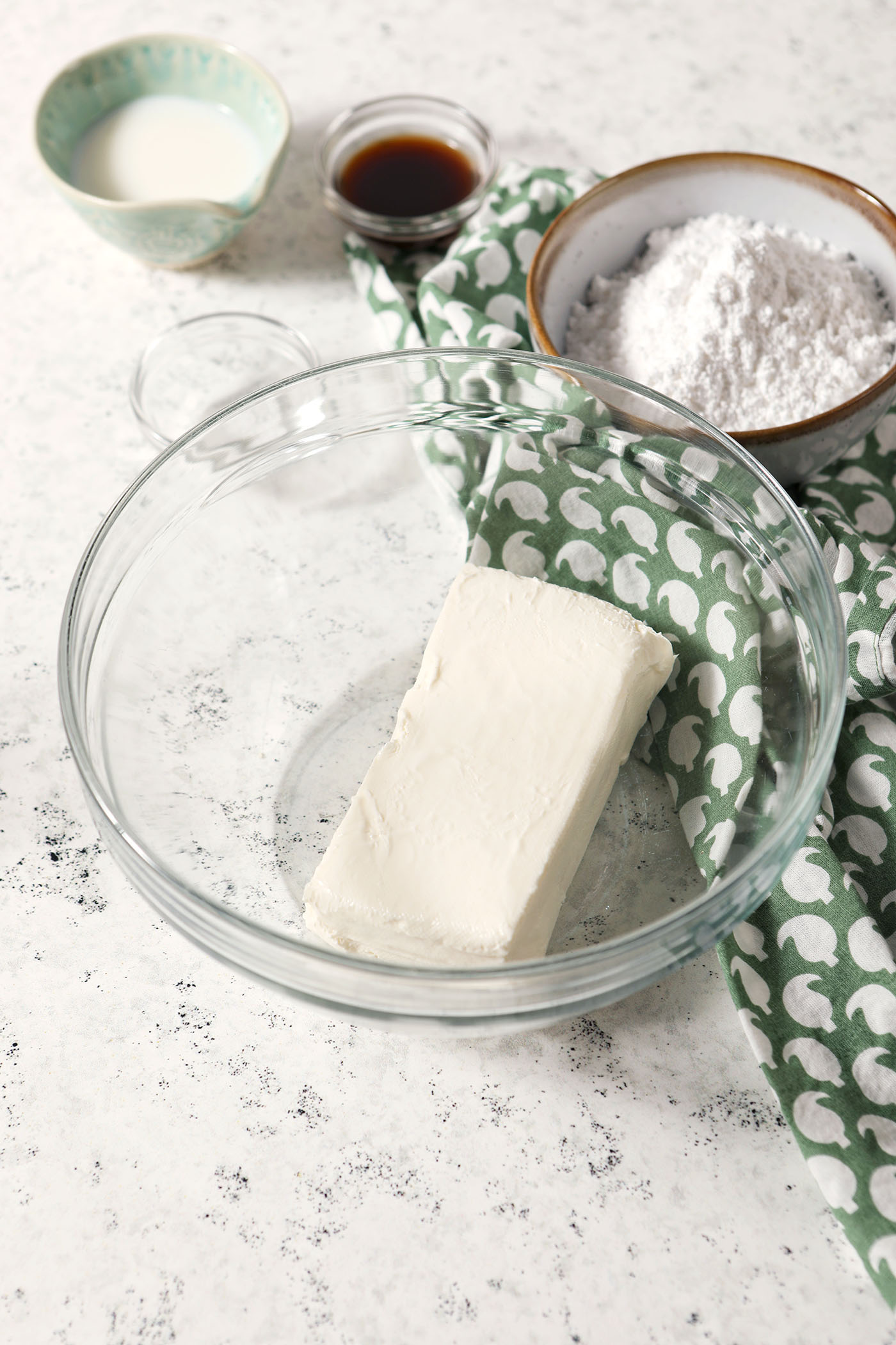 A block of cream cheese in a bowl, surrounded by bowls of other cheesecake dip ingredients on a stone surface with a green patterned linen