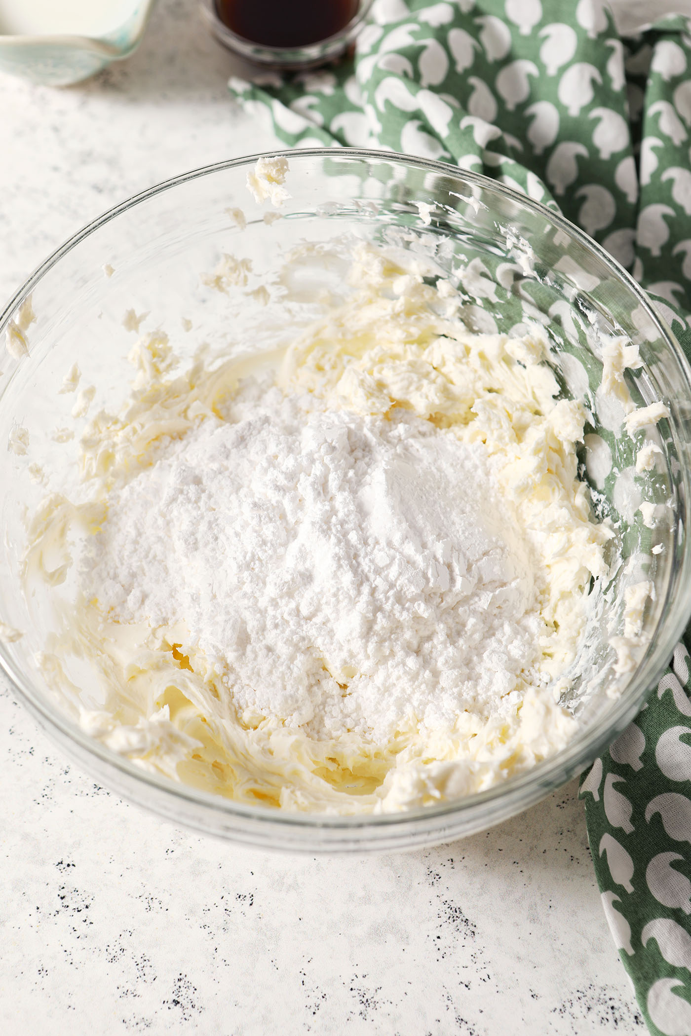 Powdered sugar sits on top of whipped cream cheese in a bowl on a stone surface next to a green patterned linen
