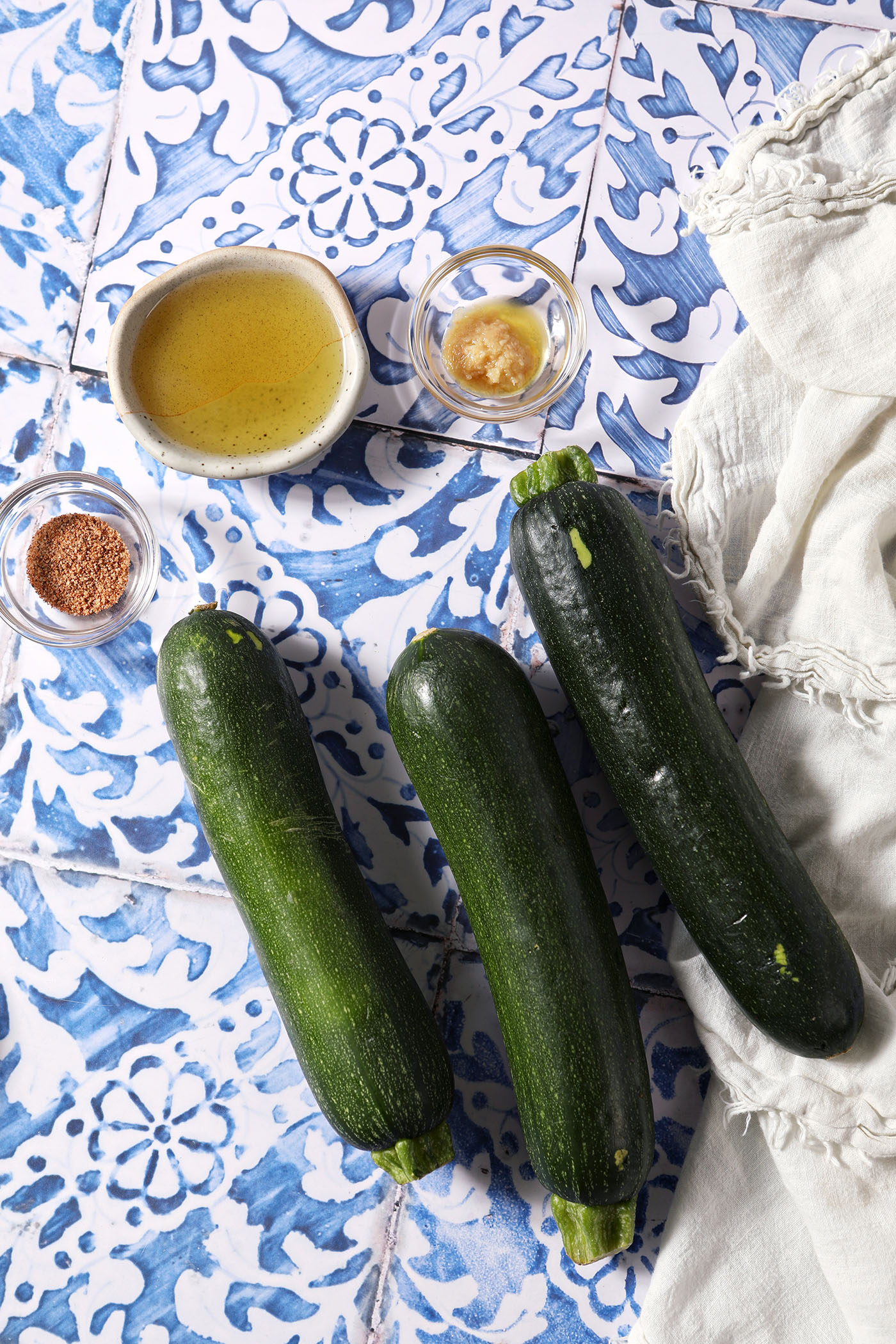 Three zucchini on a blue and white tiled surface with bowls of oil, grated garlic and Cajun seasoning.