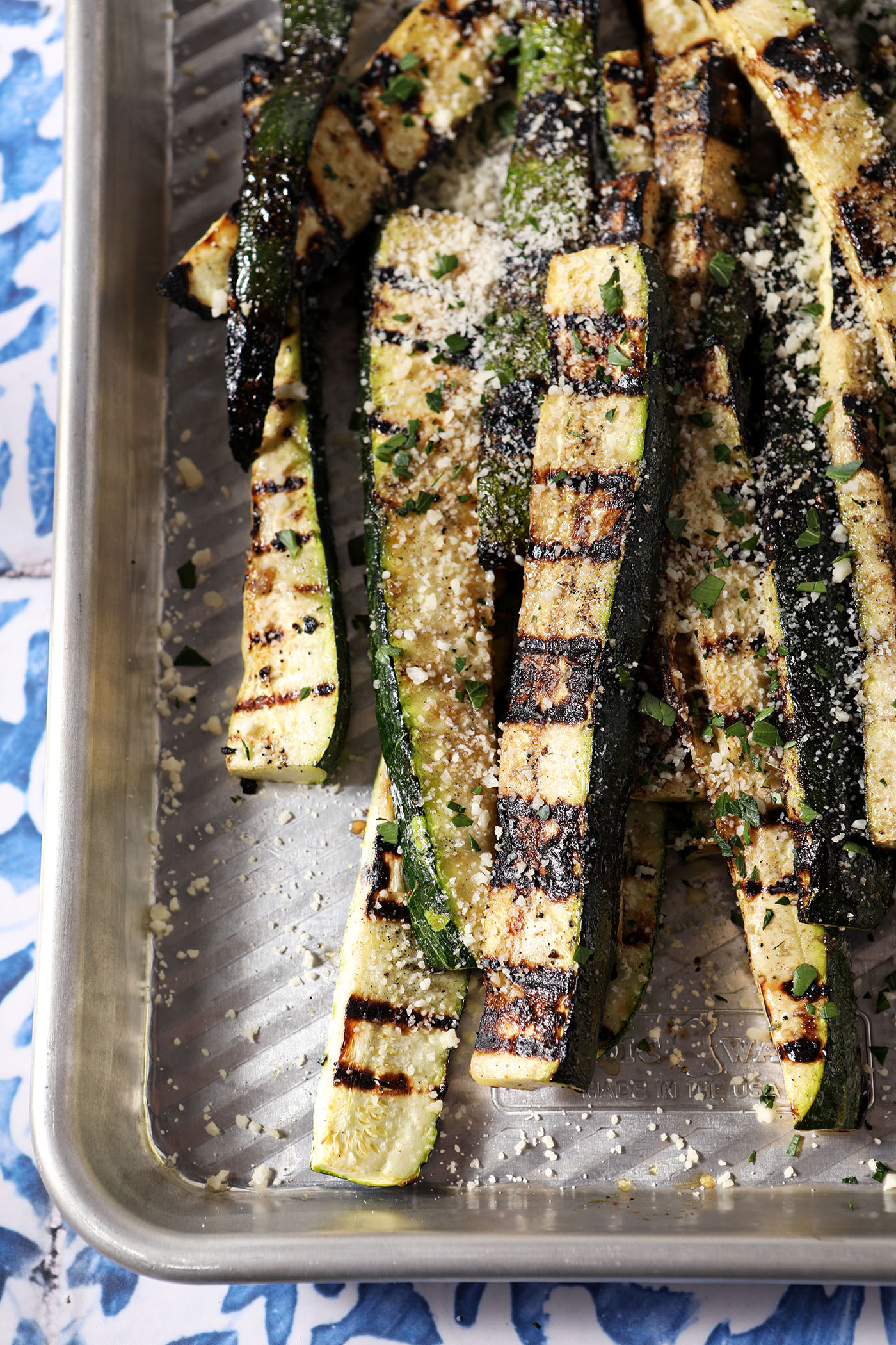 Close-up of grilled zucchini slices garnished with grated Parmesan and parsley on a silver sheet pan.