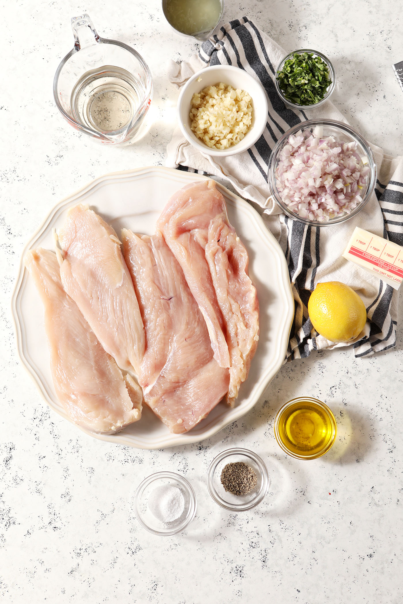 Have chicken breasts with other ingredients to make lemony chicken on a white stone surface with a black and white striped linen
