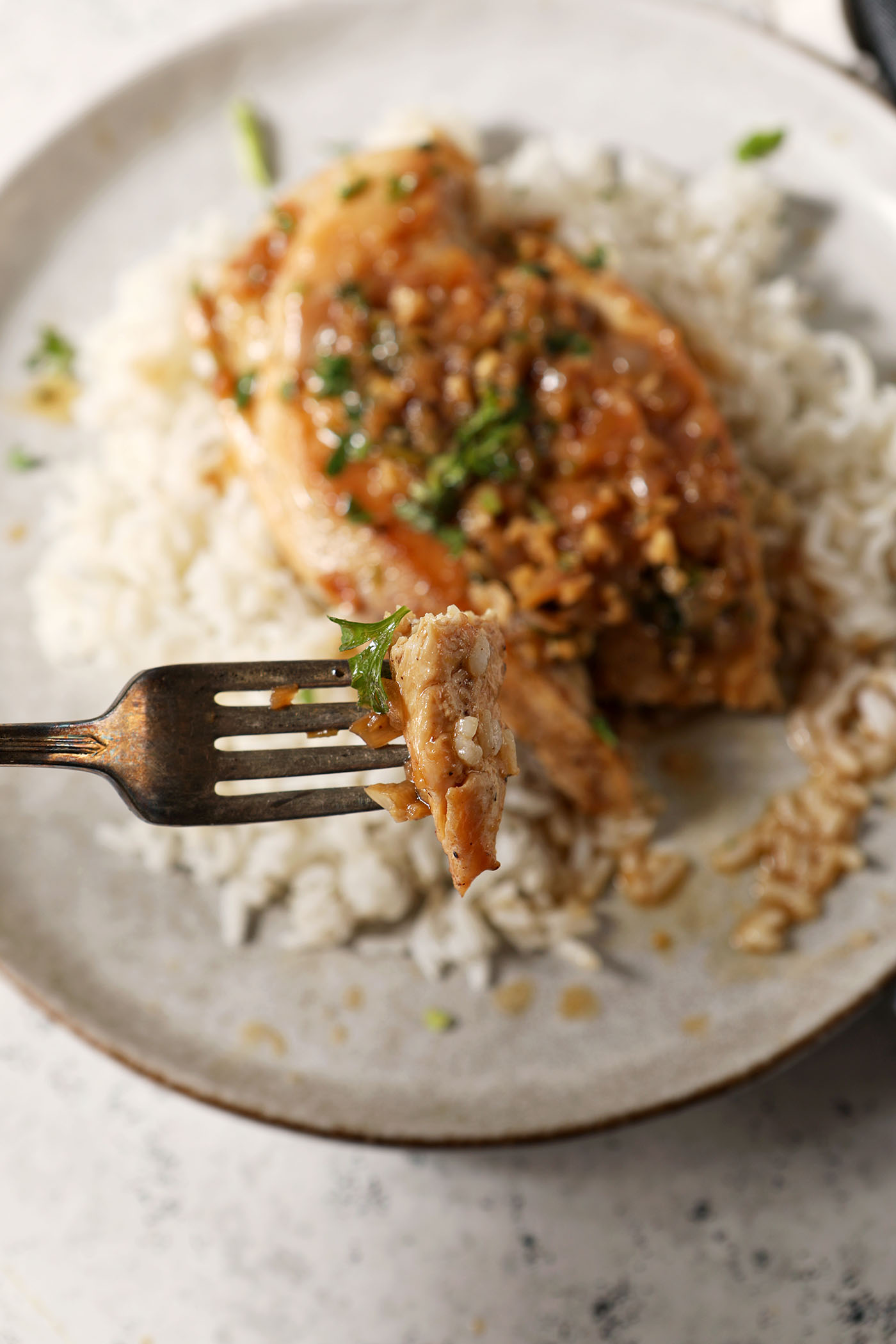 A fork holds a bite of lemon chicken above a grey plate holding a chicken breast, rice and sauce