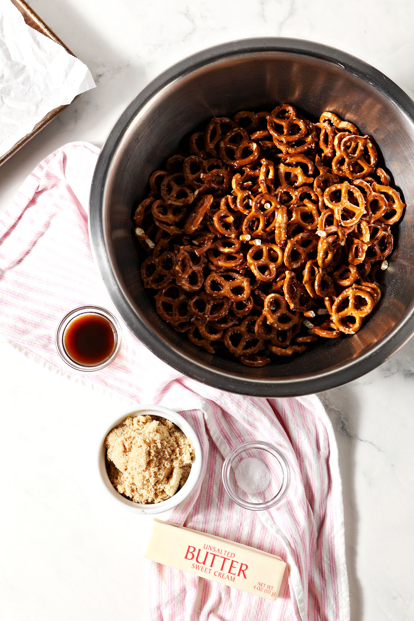 Crispy pretzels in a bowl with bowls of brown sugar, salt, vanilla, and butter around it on a stone surface with a pink and white striped lemon