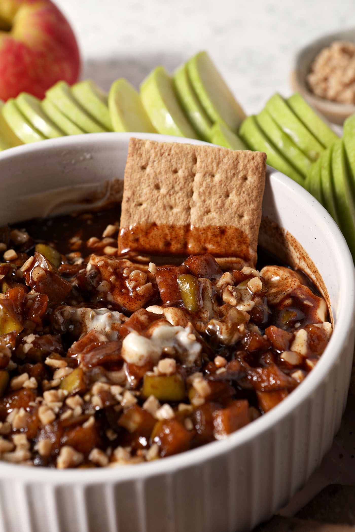A graham cracker in Caramel Apple Cheesecake Dip in a white serving dish with different dippers around it.