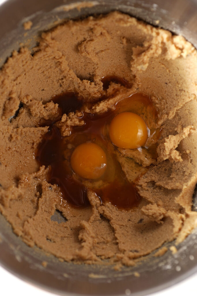 Two broken eggs and vanilla extract sit on top of creamed butter and sugar in a metal bowl