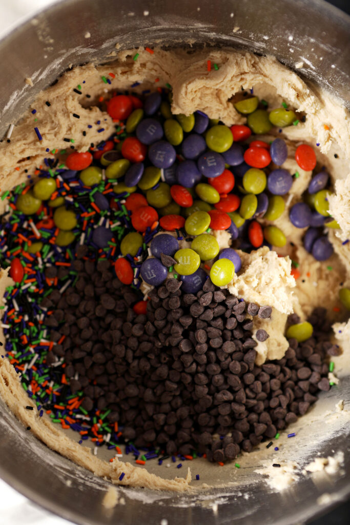 Mini chocolate chips, sprinkles, and Halloween M&M candies sit on top of cookie dough before mixing