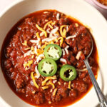A bowl of spicy bison chili from above with a spoon, garnished with shredded cheese and jalapeño