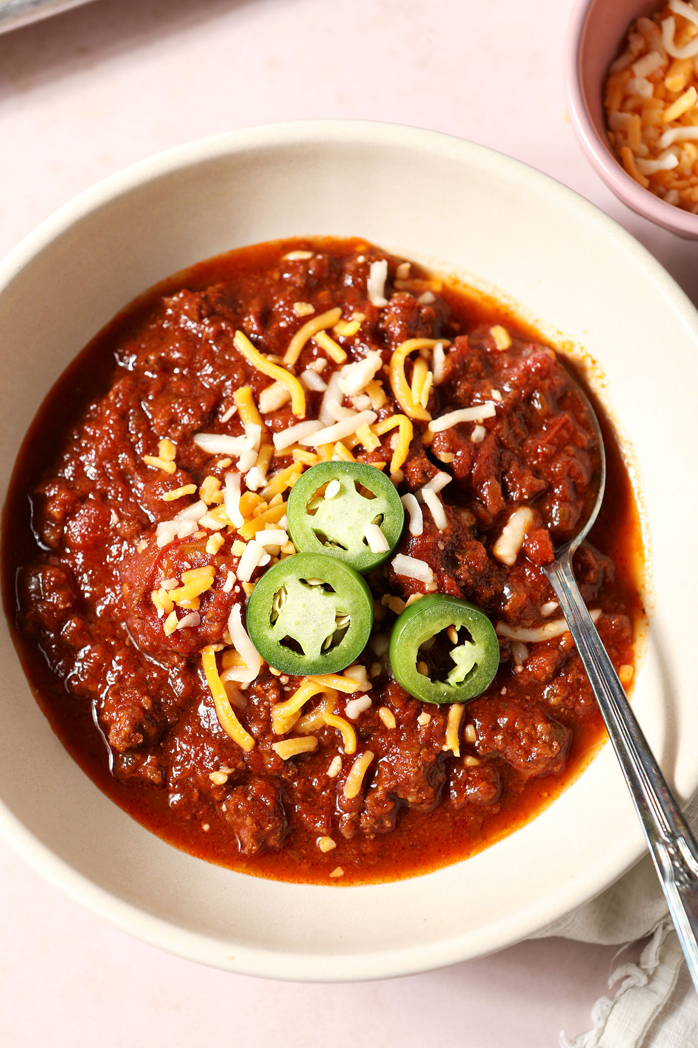 A bowl of spicy bison chili from above with a spoon, garnished with shredded cheese and jalape&ntilde;o