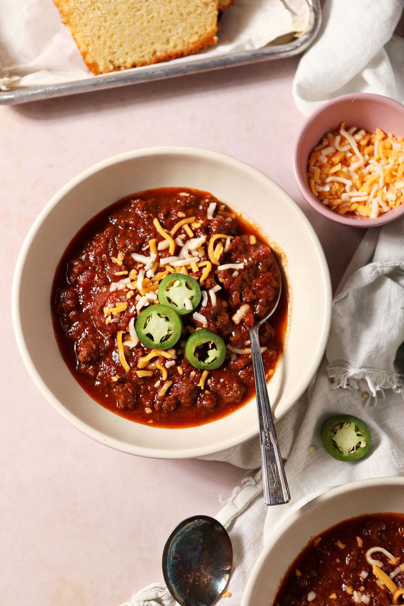 Overhead of two bowls of bison chili, garnished with shredded cheese and sliced jalapenos, on a tan surface next to a loaf of cornbread and a bowl of more cheese