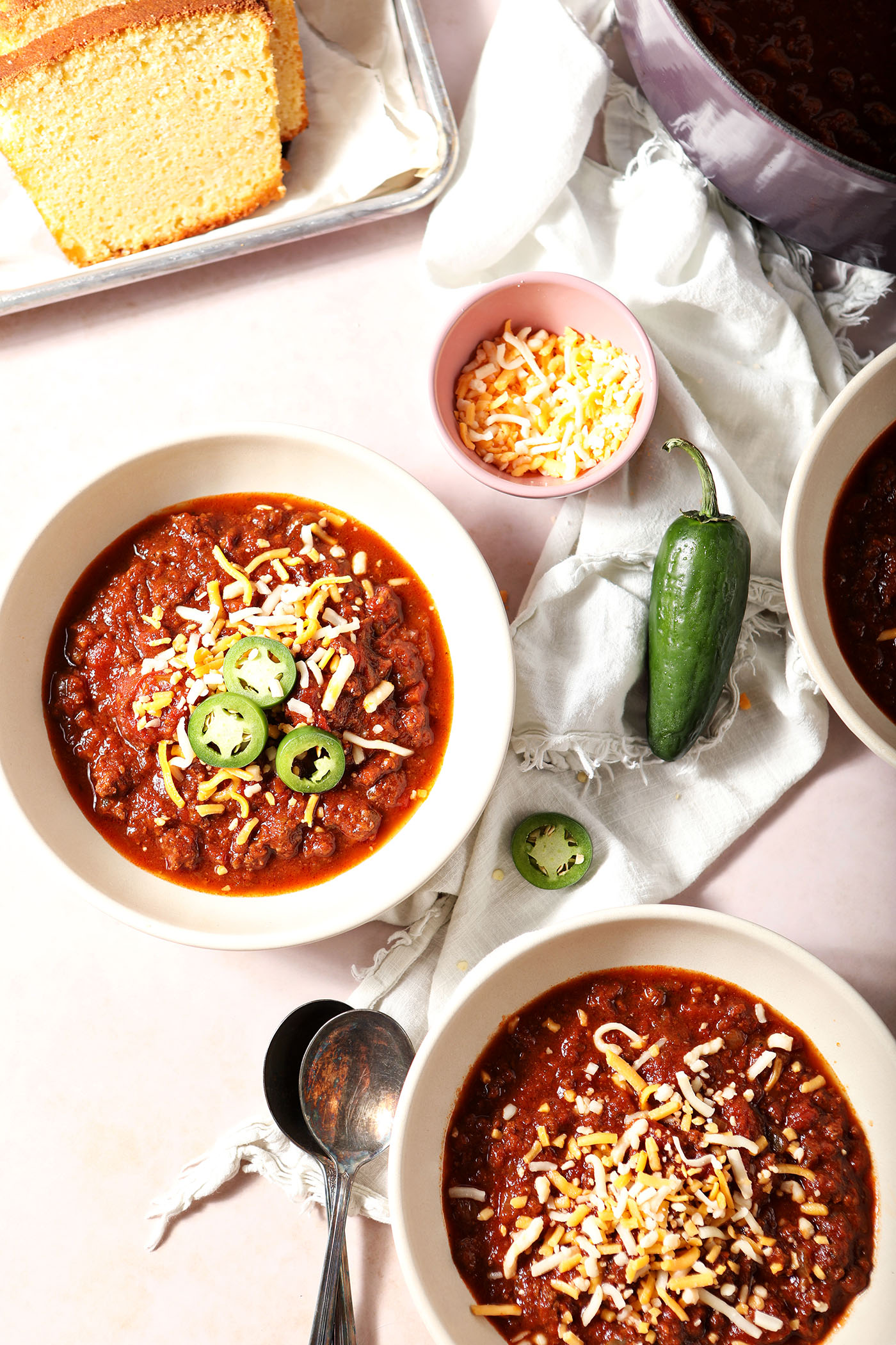 Overhead image of two bowls holding bison chili, garnished with cheese and sliced jalapenos, on a tan service, surrounded by other garnishes and a loaf of cornbread