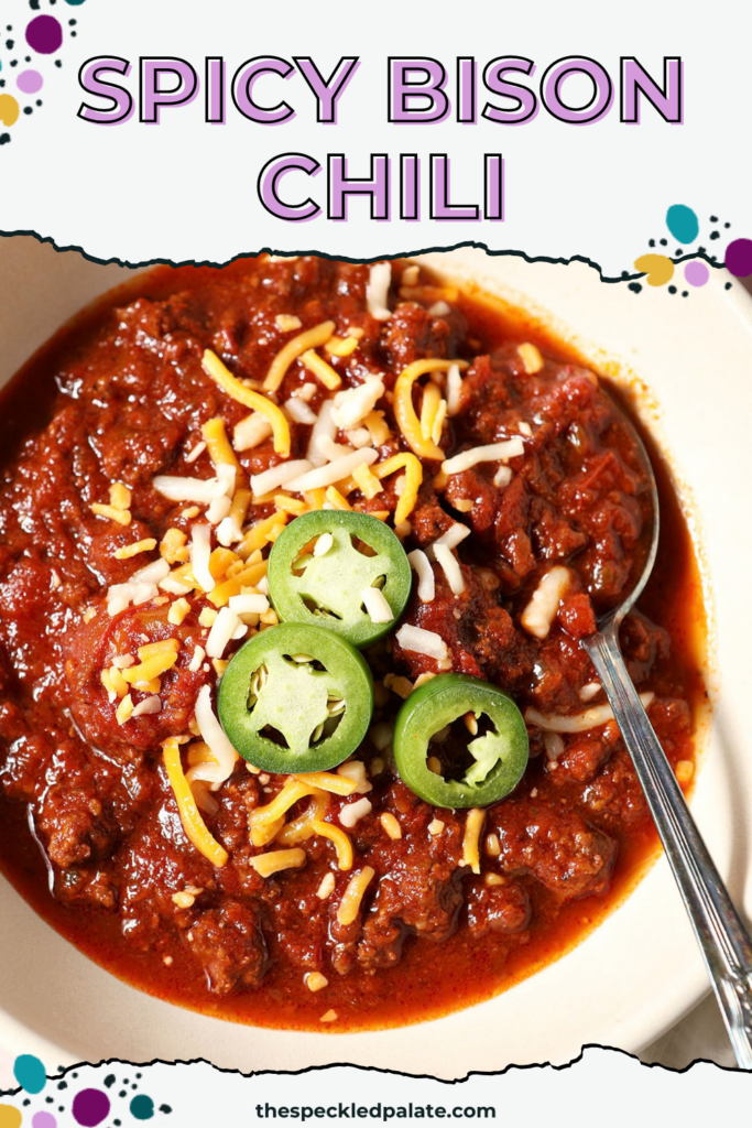 A bowl of spicy bison chili from above with a spoon, garnished with shredded cheese and jalapeño with the Pinterest Title, "Spicy Bison Chili" on top
