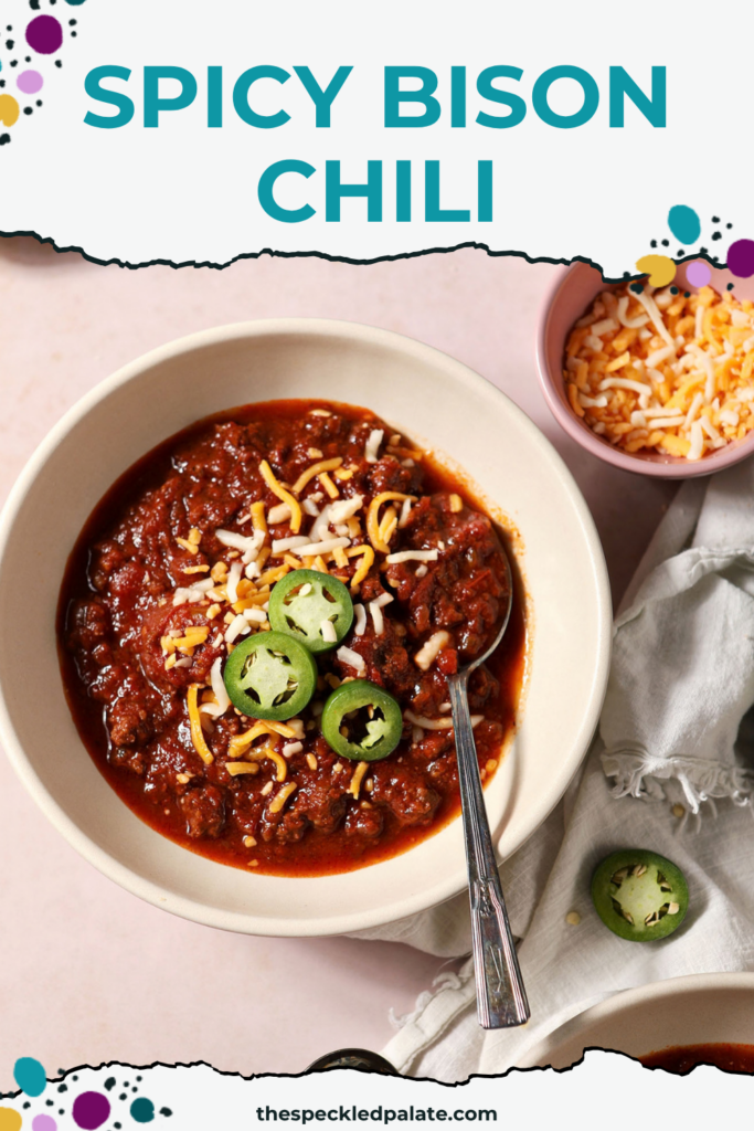 Overhead of two bowls of bison chili, garnished with shredded cheese and sliced jalapenos, on a tan surface next to a loaf of cornbread and a bowl of more cheese with the Pinterest Title, "Spicy Bison Chili" on top.
