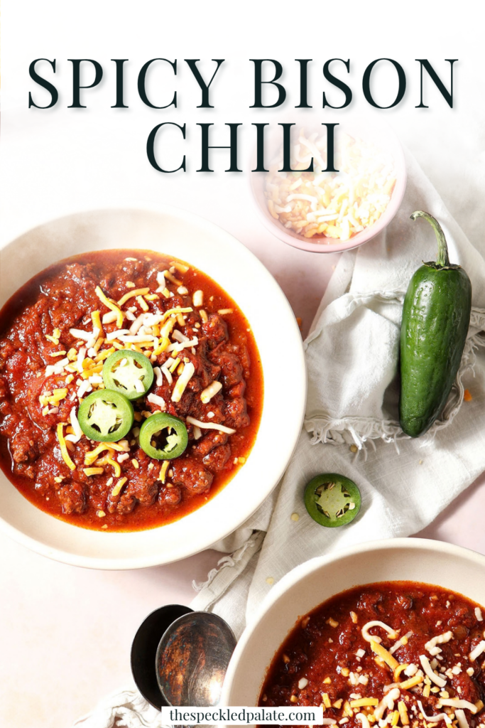 Overhead image of two bowls holding bison chili, garnished with cheese and sliced jalapenos, on a tan service, surrounded by other garnishes and a loaf of cornbread with the Pinterest Title, "Spicy Bison Chili" on top.
