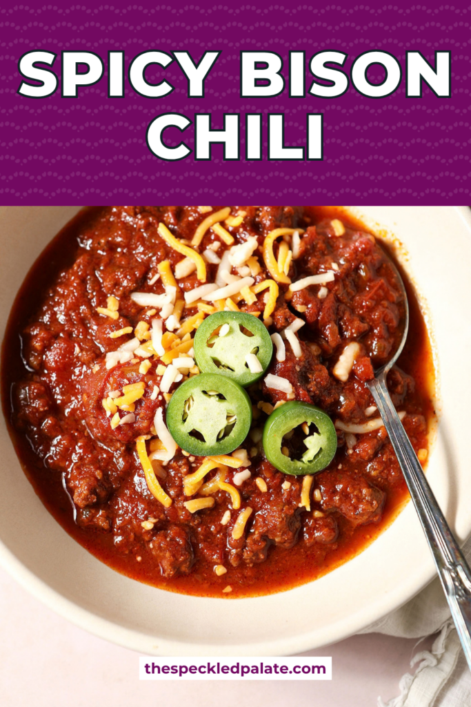A bowl of spicy bison chili from above with a spoon, garnished with shredded cheese and jalapeño with the Pinterest Title, "Spicy Bison Chili" on top